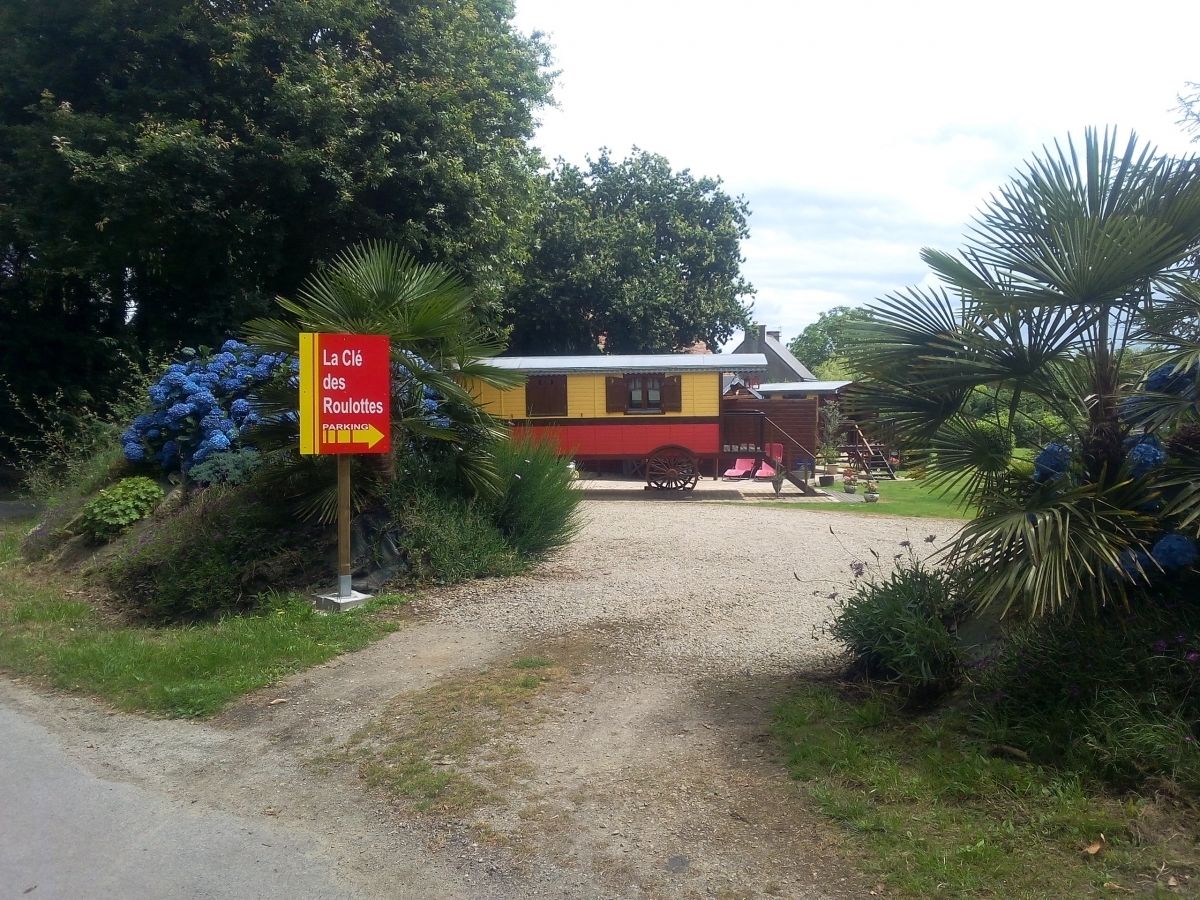 A gravel path leading to a yellow building with a red roof, surrounded by lush greenery and a sign indicating parking to the left.