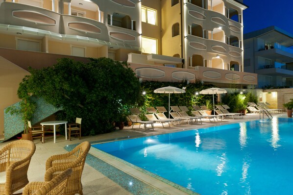 Piscina con sillas de descanso y sombrillas, rodeada de vegetación y un edificio con balcones.