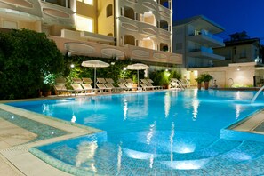 A swimming pool with lounge chairs and umbrellas, surrounded by buildings with balconies and greenery. - Hotel Derby Exclusive (Cervia)