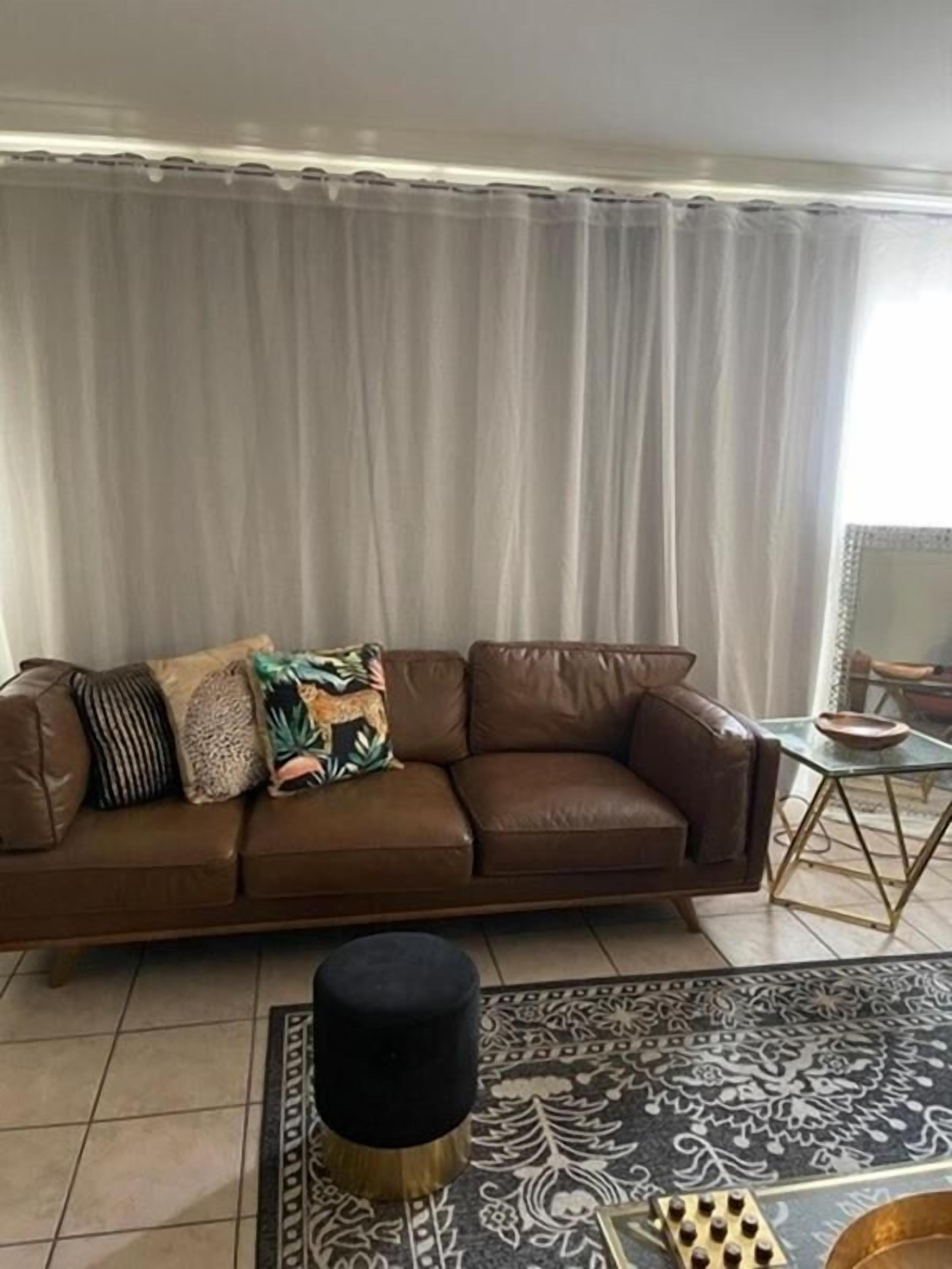 A brown leather sofa with patterned cushions, a black ottoman, and a patterned rug in a living room with a curtain backdrop.