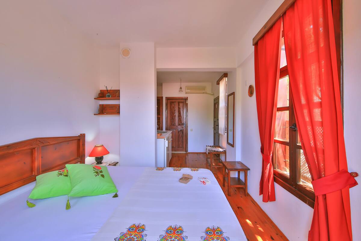 Un dormitorio con cama, mesita de noche, lámpara, ventana con cortinas rojas y vista a la cocina.