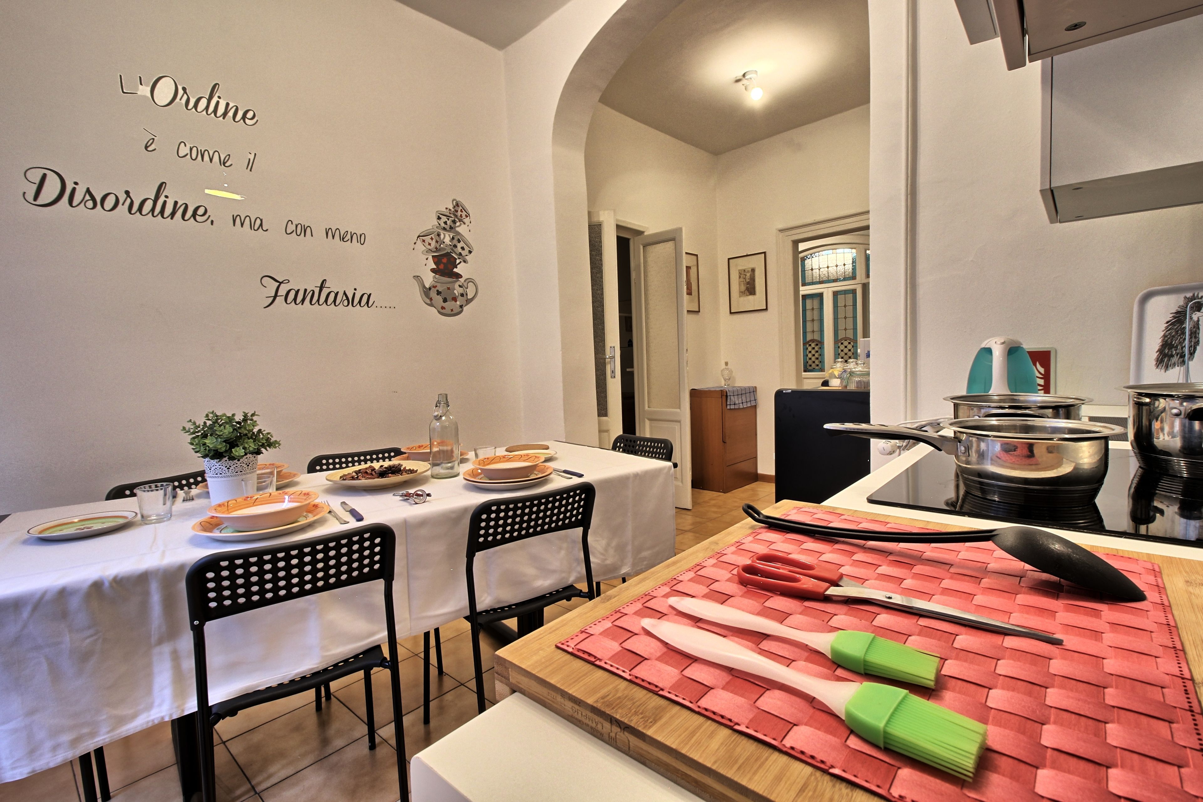 A dining area with a table set for two, chairs, a kitchen area with pots and pans, and a decorative wall quote.
