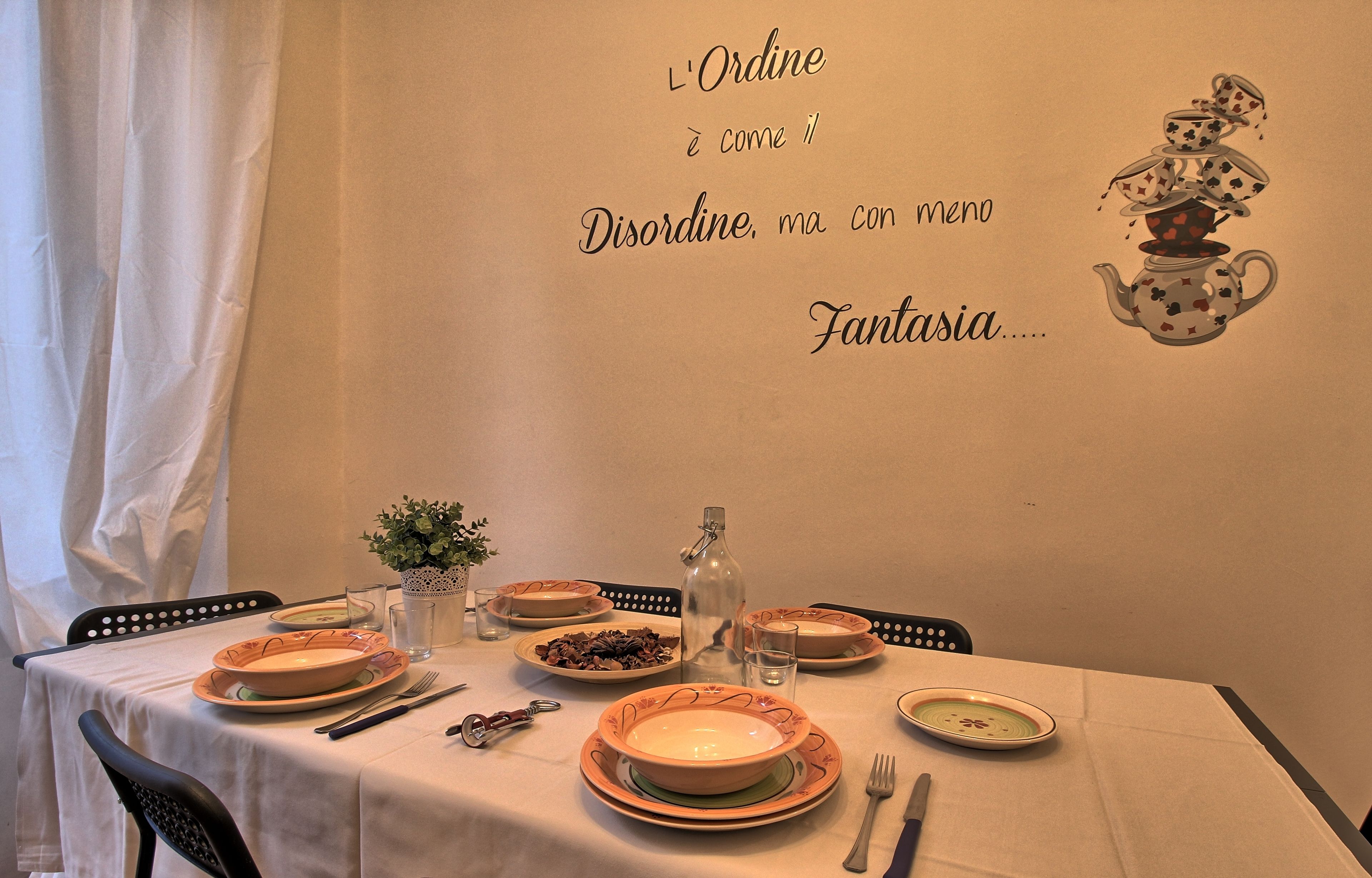 Una mesa de comedor servida con platos, tazones, cubiertos y un vaso de agua. Se observa una pared decorativa con una cita y una ilustración de una tetera.