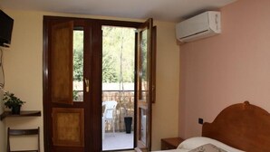 Une chambre à coucher comprenant un lit, une tête de lit en bois, une table de chevet, un climatiseur mural, une porte en bois donnant sur un espace extérieur et une petite étagère avec une plante.