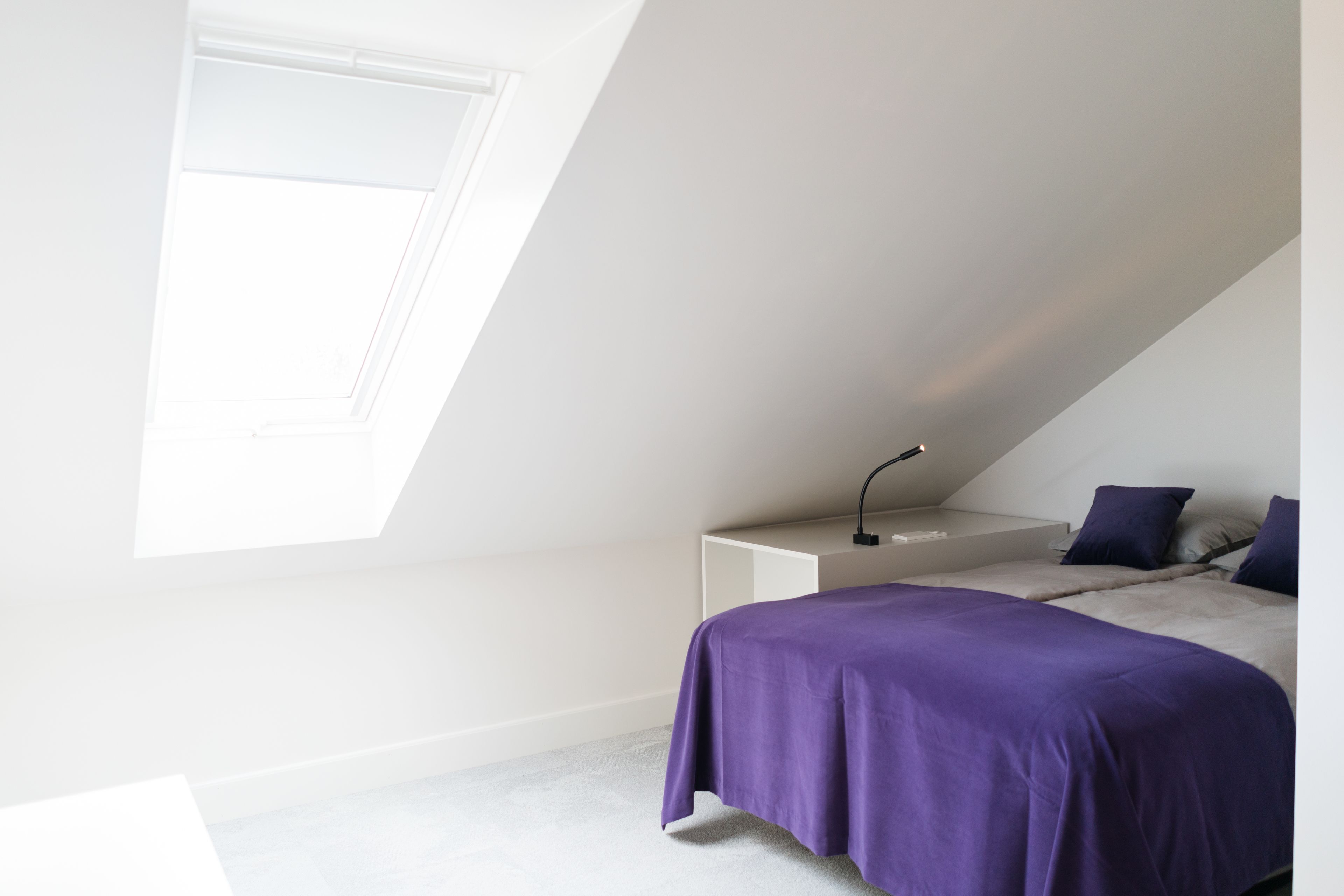 A bedroom with a sloped ceiling, a bed with a purple bedspread, a bedside table with a lamp, and a window providing natural light.