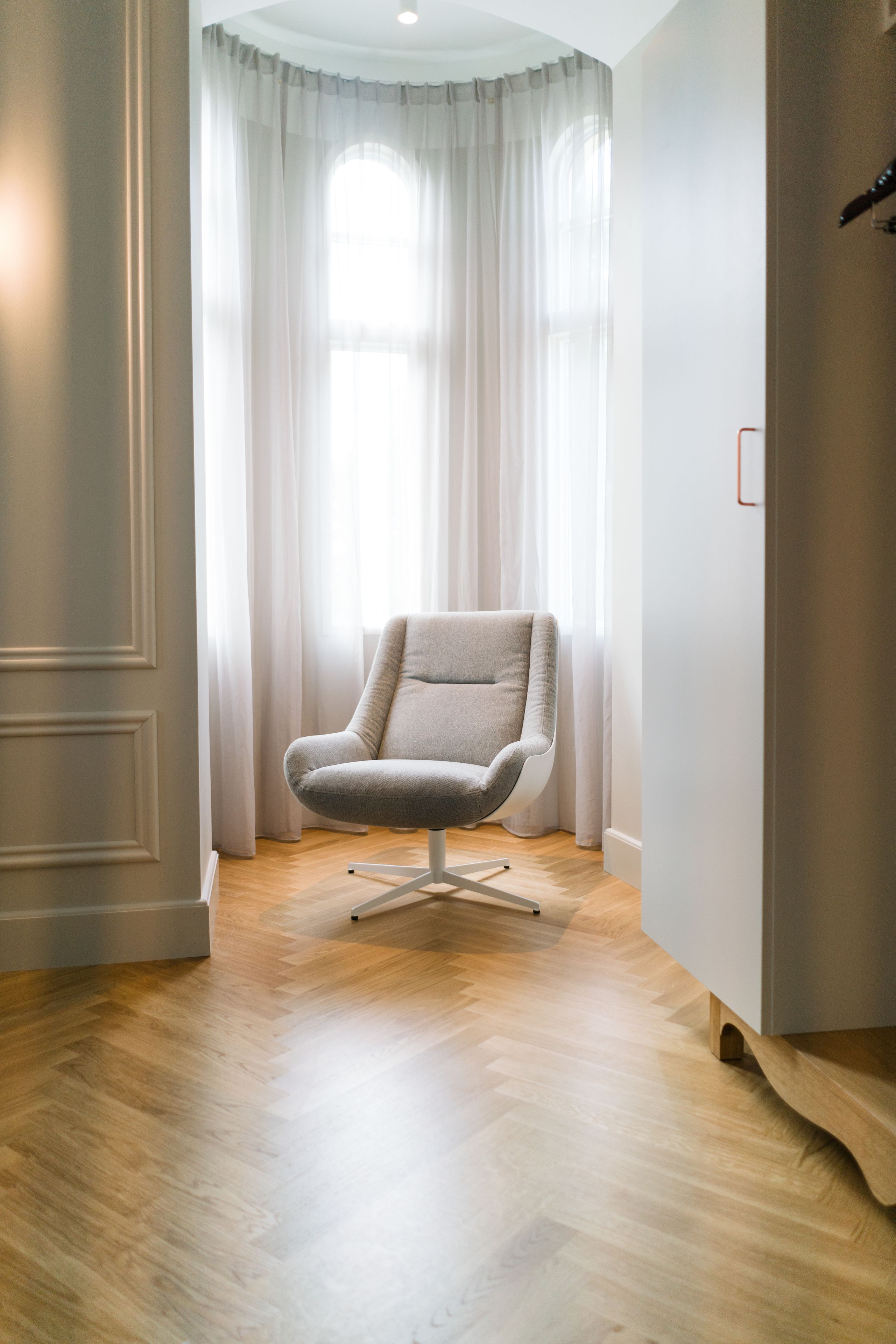Una habitación moderna con un sillón de color claro, suelo de madera y un ventanal con cortinas translúcidas.