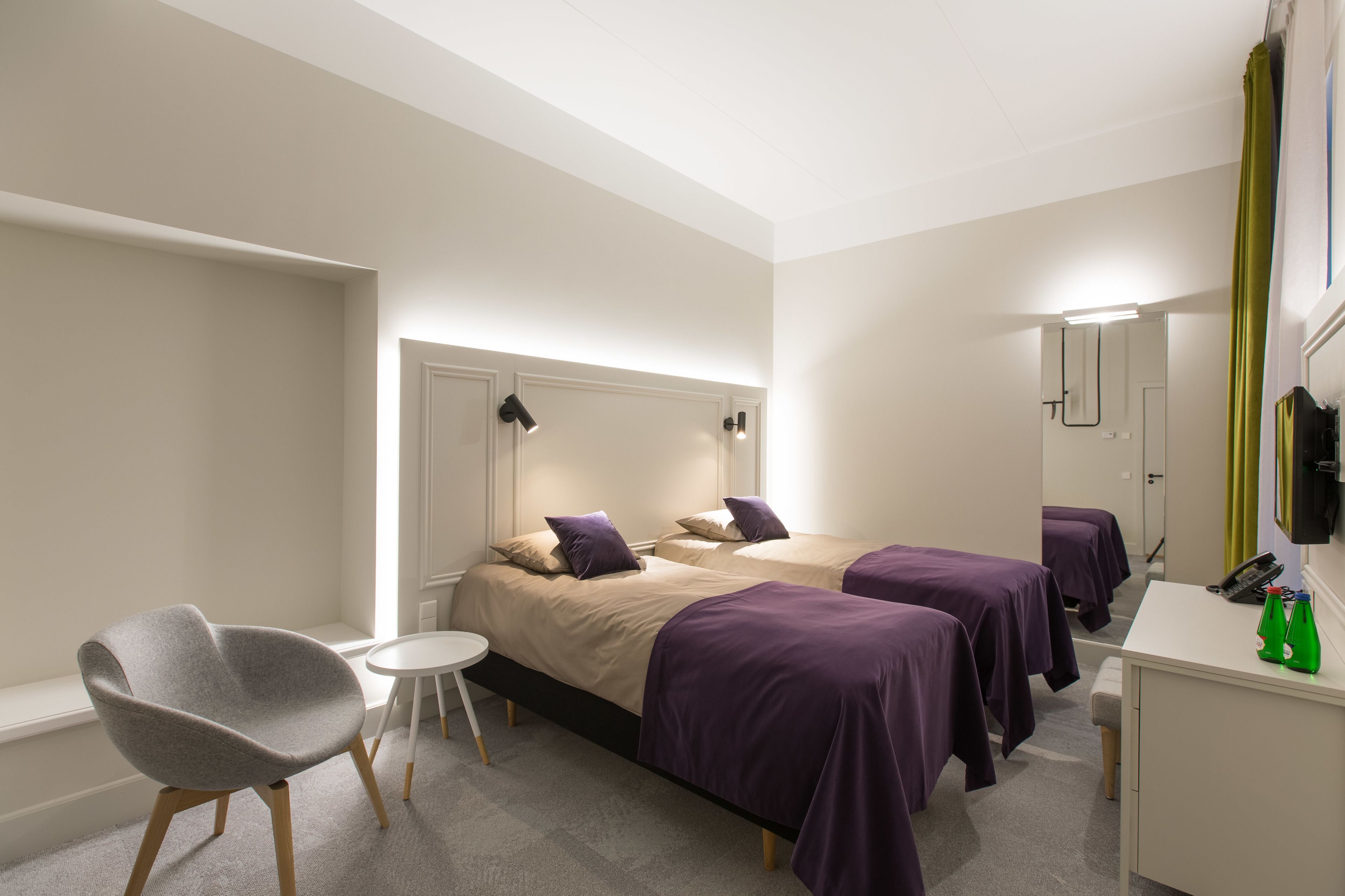 Habitación de hotel con dos camas, una silla, una mesita pequeña y un escritorio con teléfono y botellas verdes.