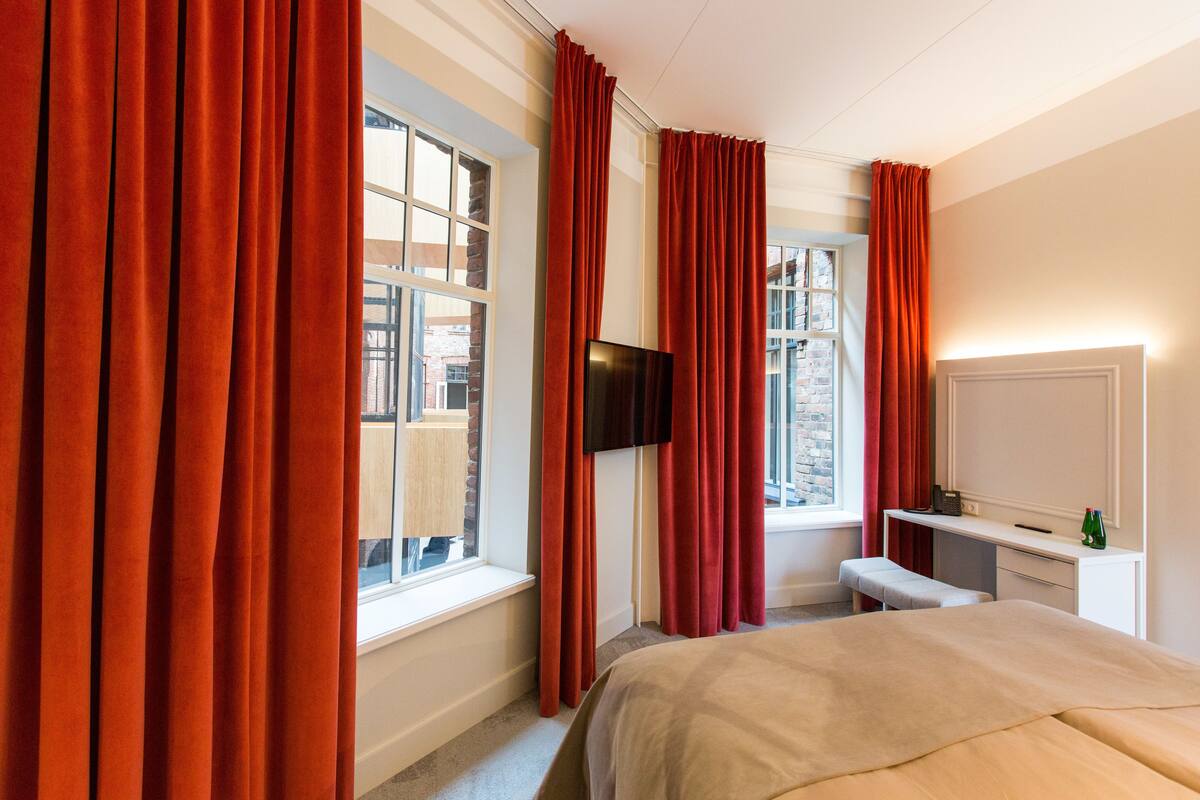 Une chambre d’hôtel avec un lit, un bureau, une télévision et de grandes fenêtres équipées de rideaux.