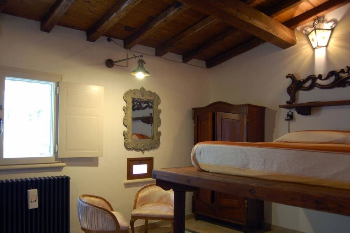 A bedroom with a bed, a chair, a table, a radiator, a window, and a wall-mounted light fixture.