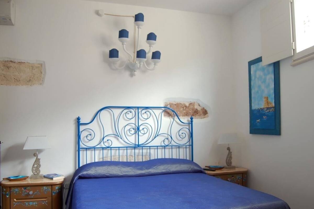 A bedroom with a blue metal bed frame, a chandelier, a painting, and a nightstand with a lamp.