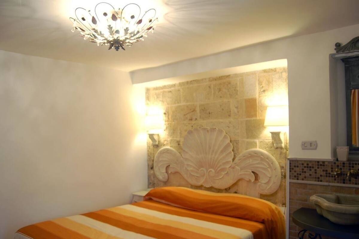 A hotel room with a bed, a decorative headboard, a wall-mounted light fixture, and a bathroom with a sink and mirror.