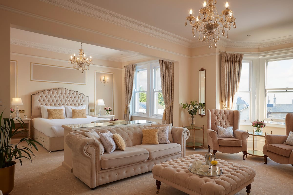 A luxurious bedroom with a large bed, a sitting area with a sofa and armchairs, a glass coffee table, and a chandelier.