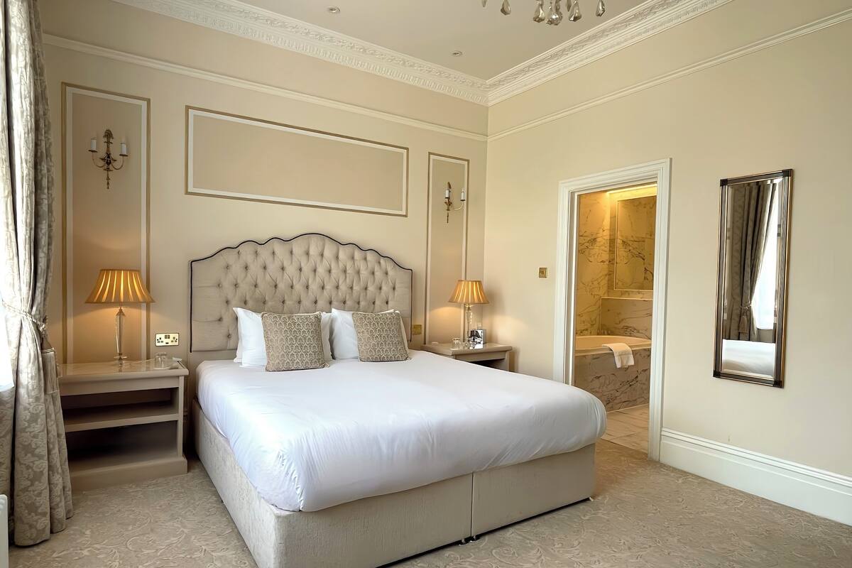 A spacious bedroom with a large bed, two bedside tables with lamps, a mirror, and a bathroom visible through an open door.