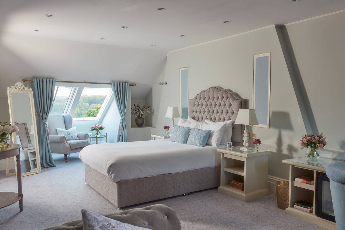 A spacious bedroom with a large bed, a sitting area with a chair and a table, and a view of the outdoors.