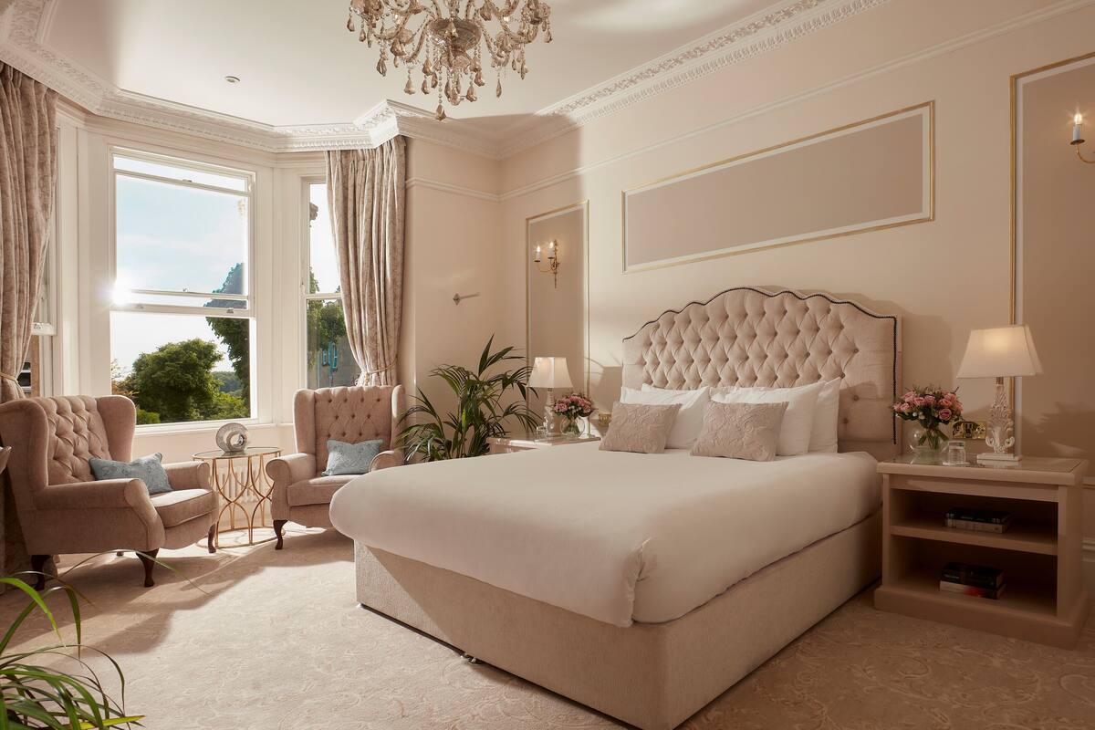 A spacious bedroom with a large bed, a sitting area with a chair and a side table, and a view of the outdoors through a window.