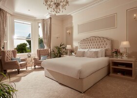A spacious bedroom with a large bed, a sitting area with a chair and a side table, and a view of the outdoors through a window.