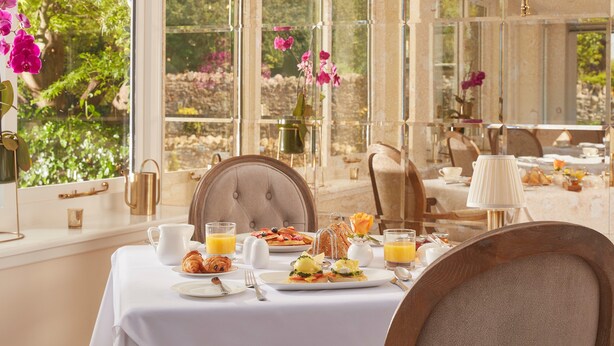 A breakfast table set with croissants, fruit, and juice, overlooking a garden with pink flowers.
