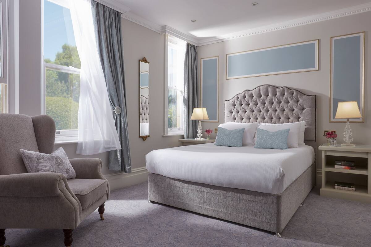 A bedroom with a large bed, a chair, a nightstand, a mirror, and a window with curtains.