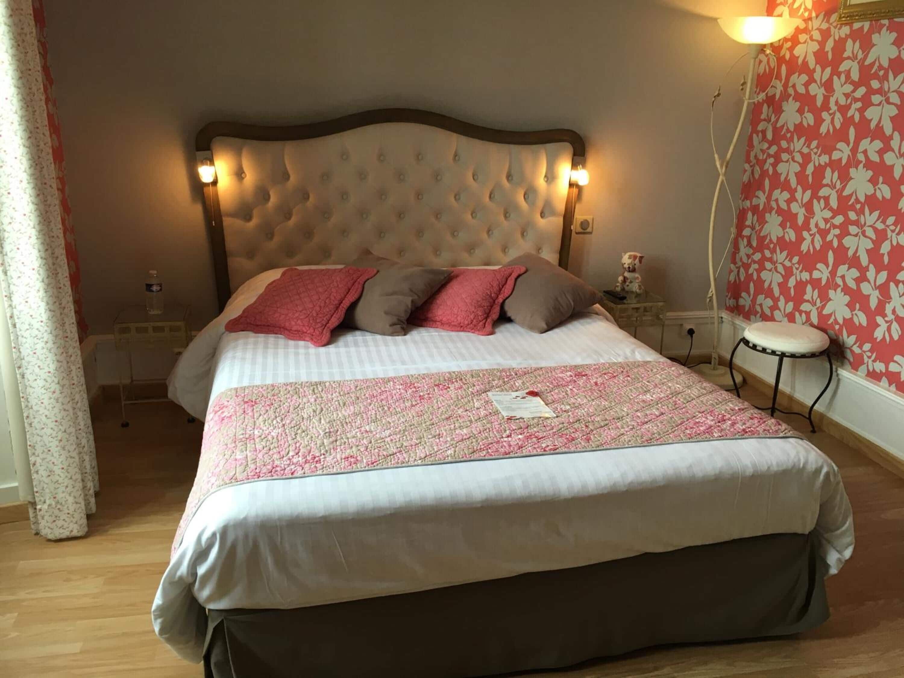 A bedroom with a large bed, a bedside table, a nightstand, a small stool, and a wall with a floral pattern.