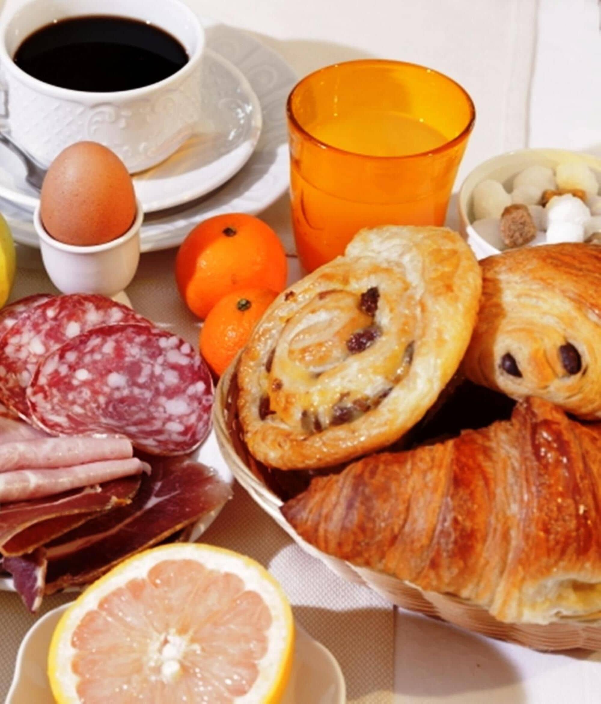 Una mesa de desayuno con pasteles, una taza de café, un vaso de zumo de naranja, un plato de embutidos y una rodaja de limón.