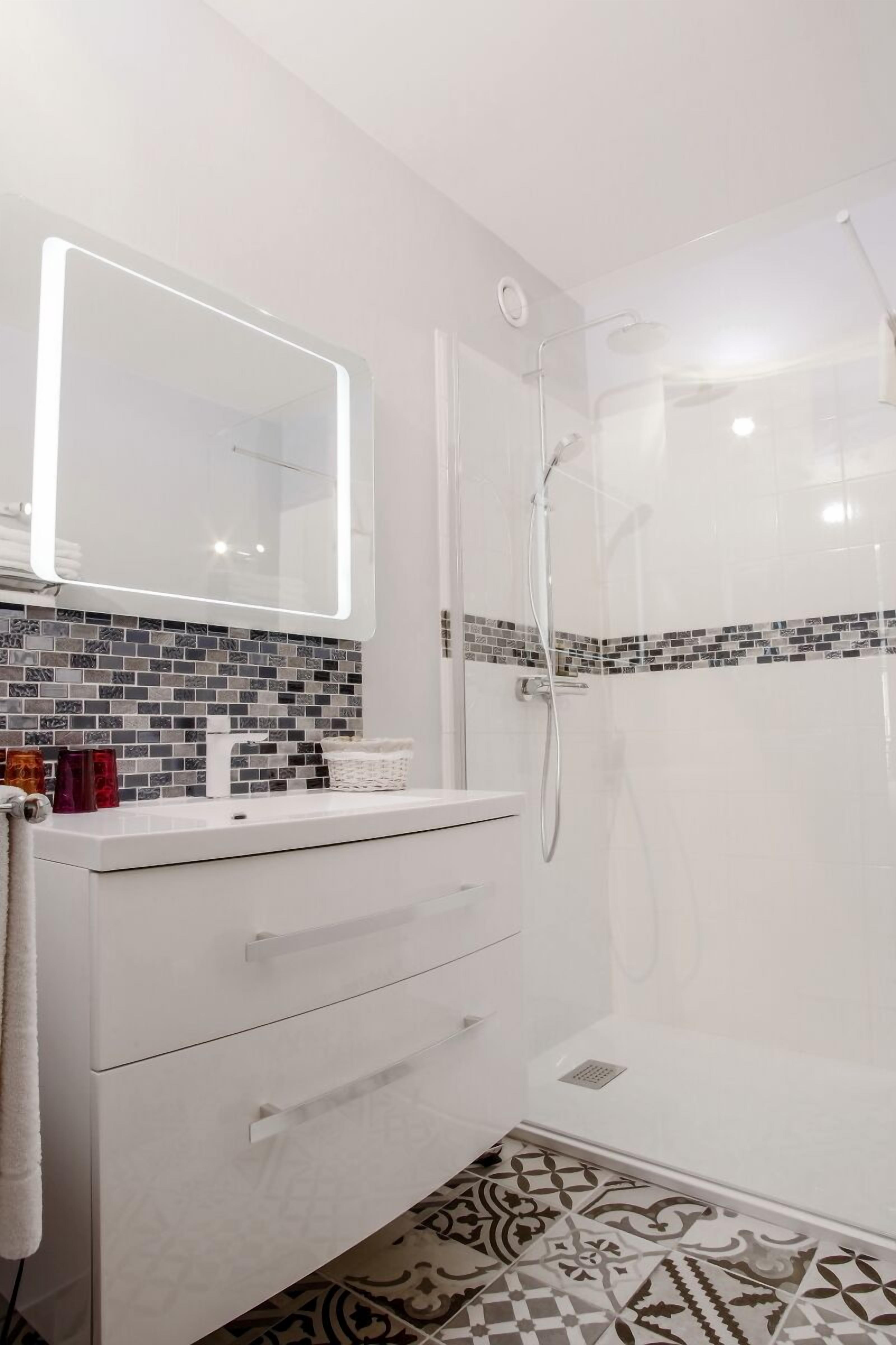 A modern bathroom with a white vanity, a large mirror with integrated lighting, a shower enclosure, and patterned floor tiles.