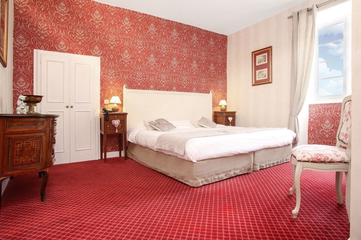 Ein Schlafzimmer mit roten Tapeten mit Muster, einem weißen Bett, einer Holzkommode und einem Stuhl.