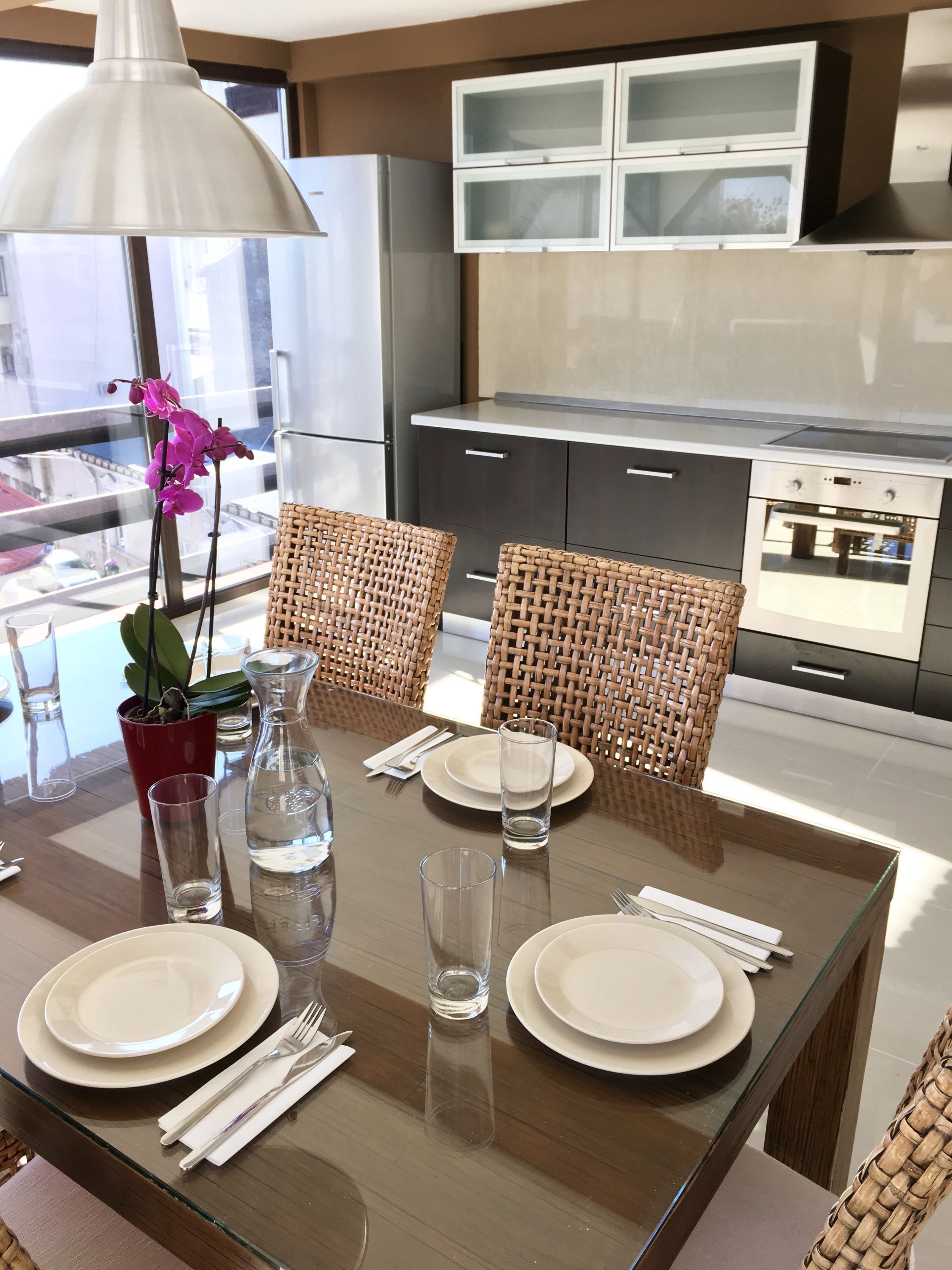 Un'area pranzo moderna con un tavolo in vetro, sedie in vimini e vista sulla cucina sullo sfondo.