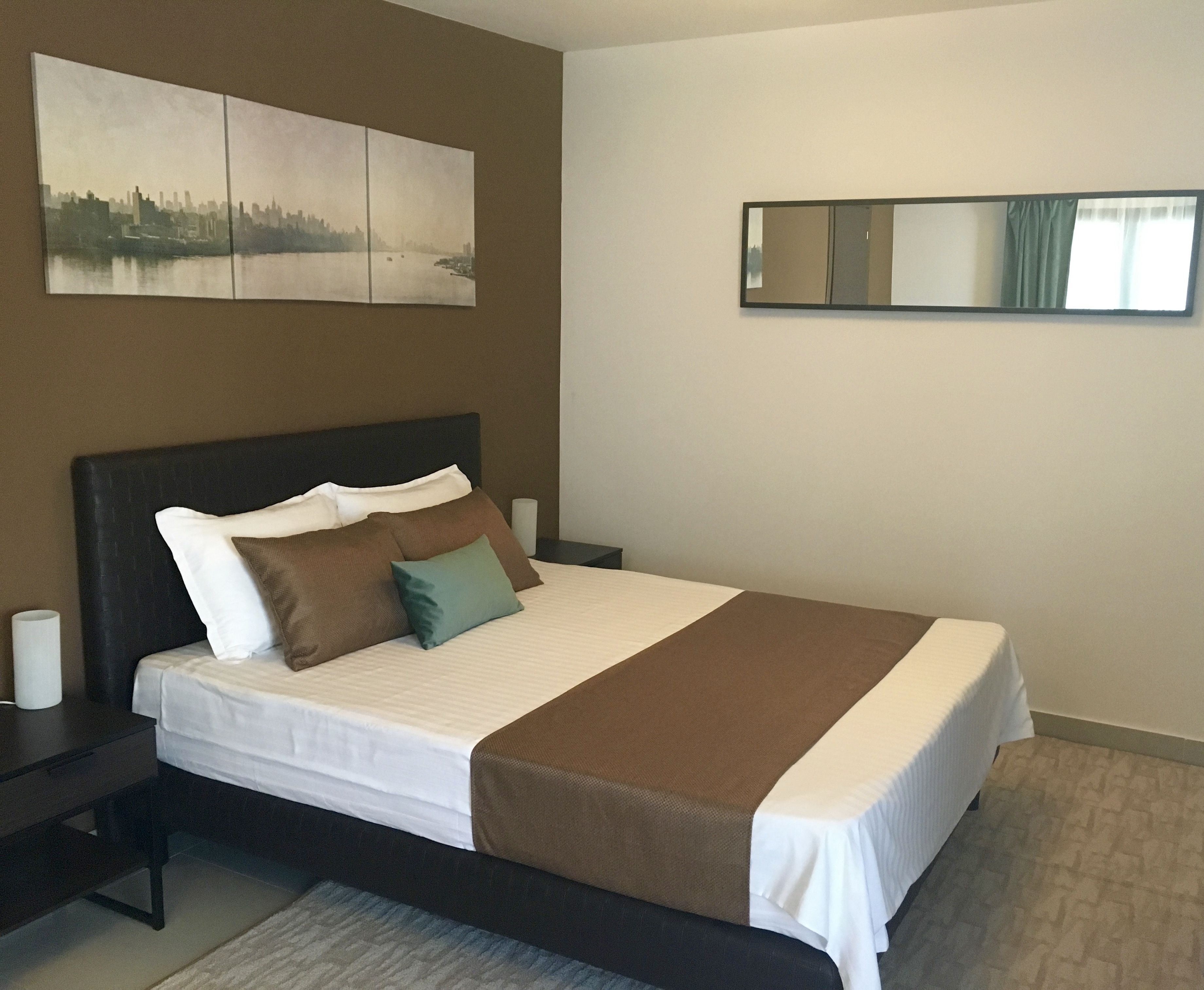 Una cama bien hecha con una manta y almohadas marrones, un cabecero oscuro y una pintura de un paisaje urbano encima.