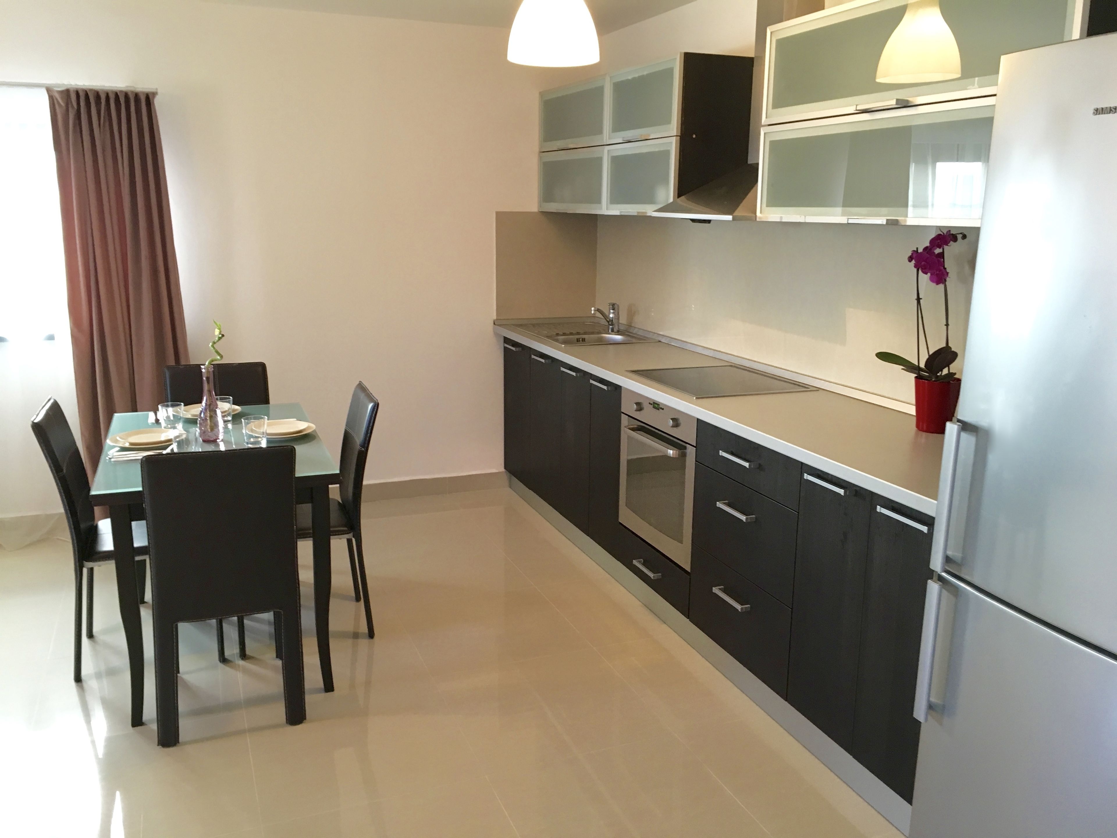 A modern kitchen with dark cabinets, a stainless steel refrigerator, a built-in oven, and a dining area with a table and chairs.