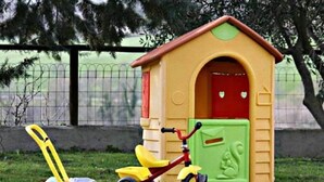 Une petite maison de jeu avec une porte rouge et un toit jaune, un tricycle rouge et une tondeuse à gazon jaune devant.
