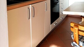 Une cuisine avec des meubles de couleur beige, un plan de travail noir, une machine à café et une table en bois recouverte d’une nappe à carreaux.