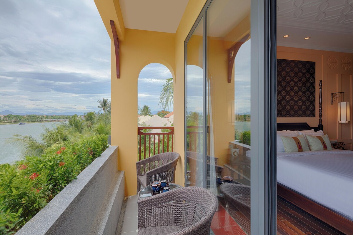 Un balcón con mobiliario de mimbre, una cama con ropa de cama blanca y vistas a una masa de agua y montañas distantes.