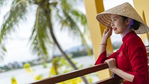 A person in traditional attire with a conical hat, seated on a bench with a view of palm trees and a body of water. - Little Riverside. A Luxury Hotel & Spa (Da Nang)