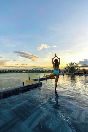 Uma pessoa fazendo uma postura de yoga no deck da piscina com vista para o pôr do sol.