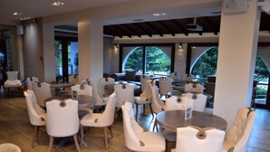 A spacious indoor dining area with round tables and white upholstered chairs, large windows with arched tops, and a wooden floor. - Konaki Hotel & SPA (Aristotelis)