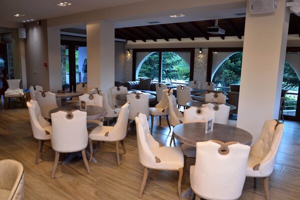 A spacious indoor dining area with round tables and white upholstered chairs, large windows with arched tops, and a wooden floor. - Konaki Hotel & SPA (Aristotelis)