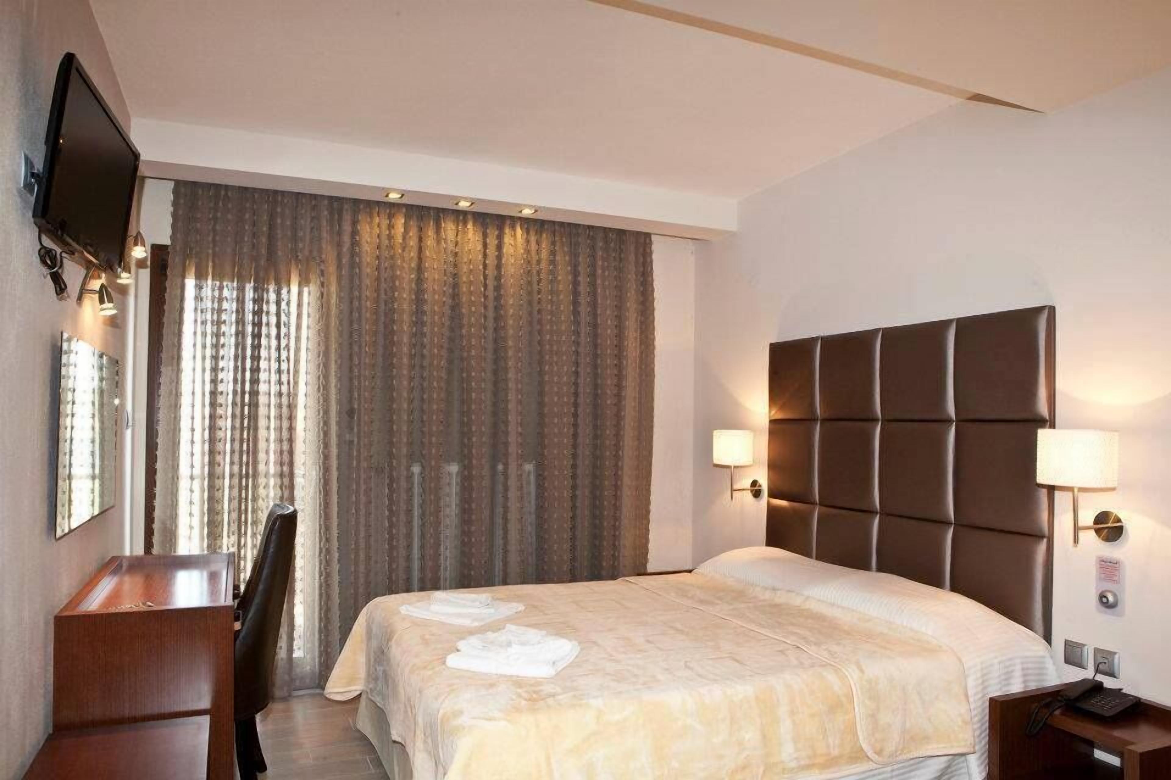 Ett hotellrum med en stor säng, ett skrivbord, en stol, en tv som är monterad på väggen och ett fönster med gardiner.
