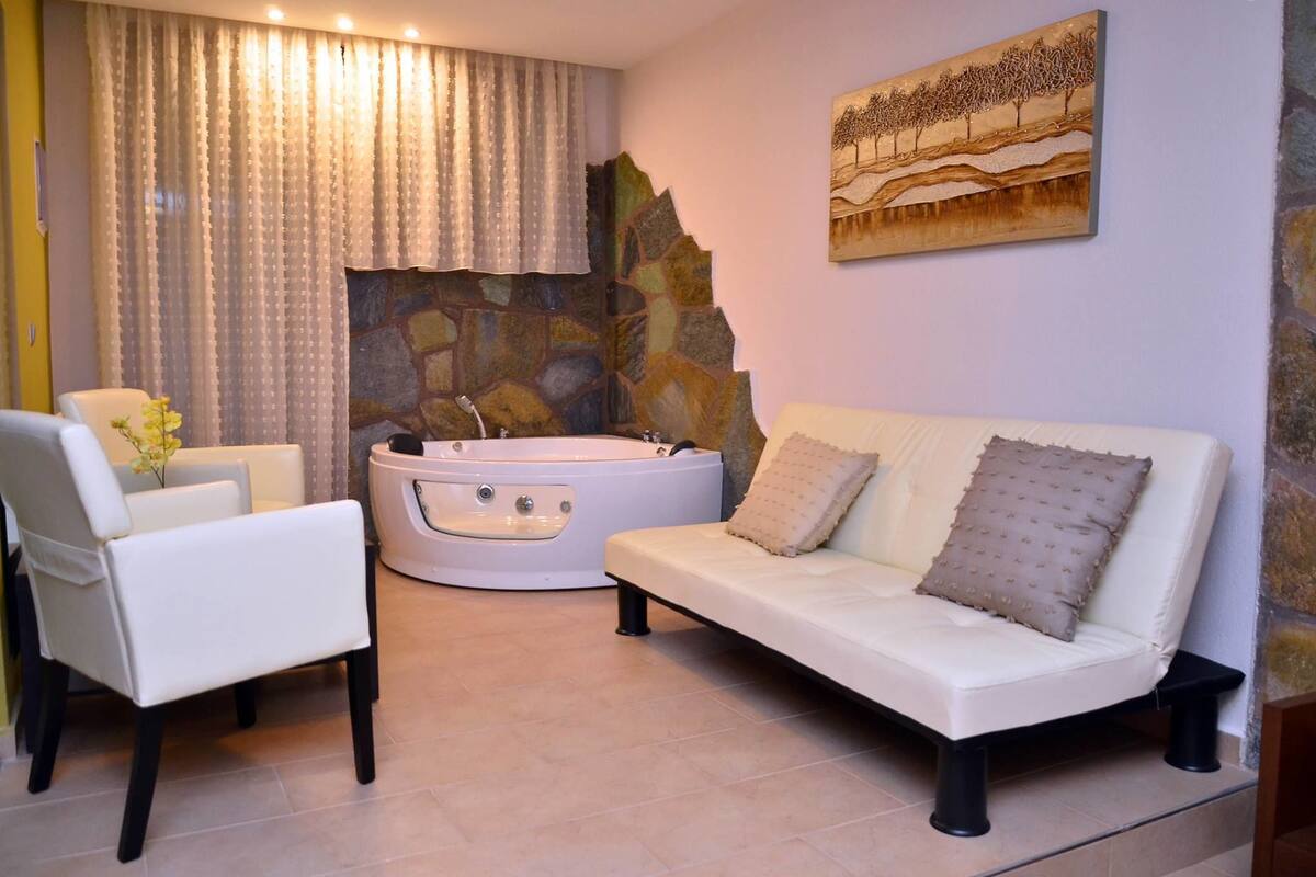 A spa room with a white sofa, a white chair, a stone wall, a painting, and a bathtub.
