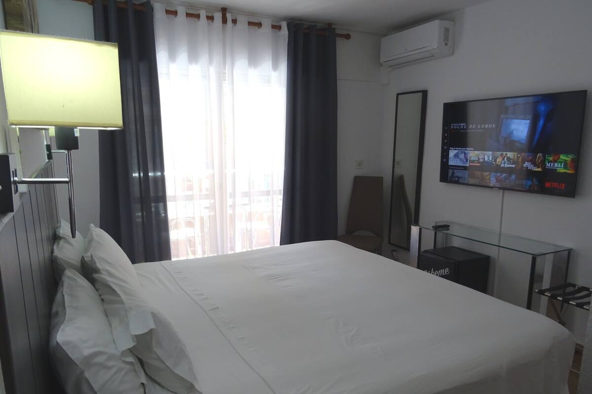 Ein Hotelzimmer mit Bett, Wandfernseher, Fenster mit Vorhängen und Klimaanlage.