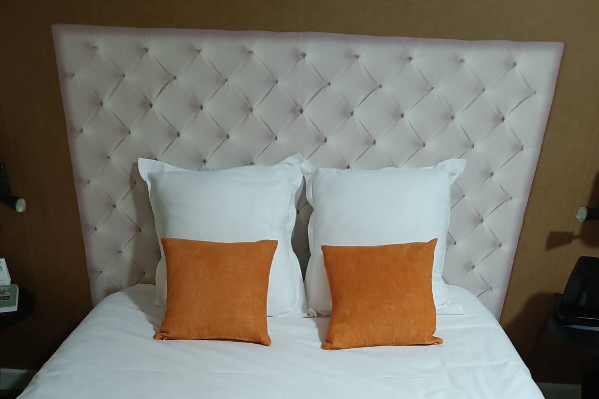 Una cama con cabecera capitonada, ropa de cama blanca y dos almohadas naranjas.