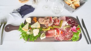 Una tabla de embutidos con diversos tipos de carne, quesos y encurtidos sobre una tabla de madera, acompañada de cubiertos y una servilleta sobre un mantel blanco.