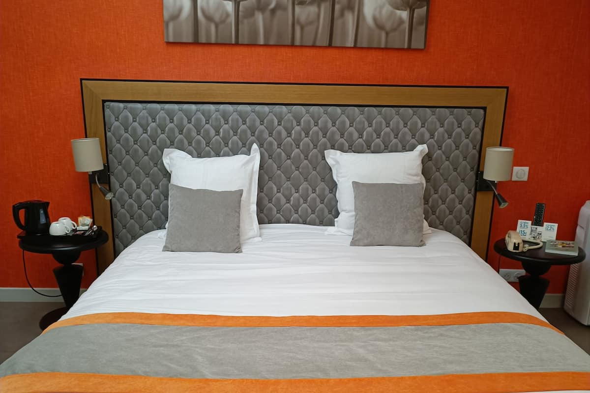 Una cama bien hecha con un cabecero gris capitonado, almohadas blancas y grises, y una colcha anaranjada y gris.