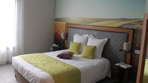 Una habitación de hotel con una cama grande, cabecera con diseño texturizado, mesita de noche con lámpara y un cuadro en la pared que muestra un paisaje.