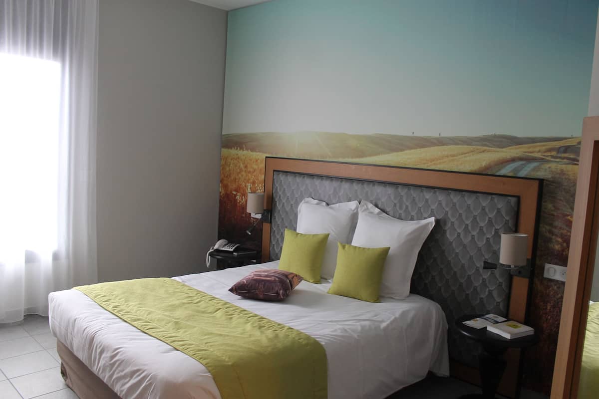 Una habitación de hotel con una cama grande, cabecera con diseño texturizado, mesita de noche con lámpara y un cuadro en la pared que muestra un paisaje.