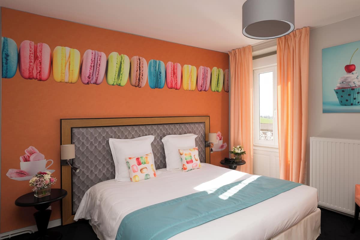 Habitación de hotel con una cama, una silla, una mesita de noche, una lámpara, una ventana con cortinas y un mural de macarons coloridos en la pared.