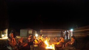 Un gruppo di persone riunite attorno a un fuoco da campo di notte, sedute a terra con tende sullo sfondo.