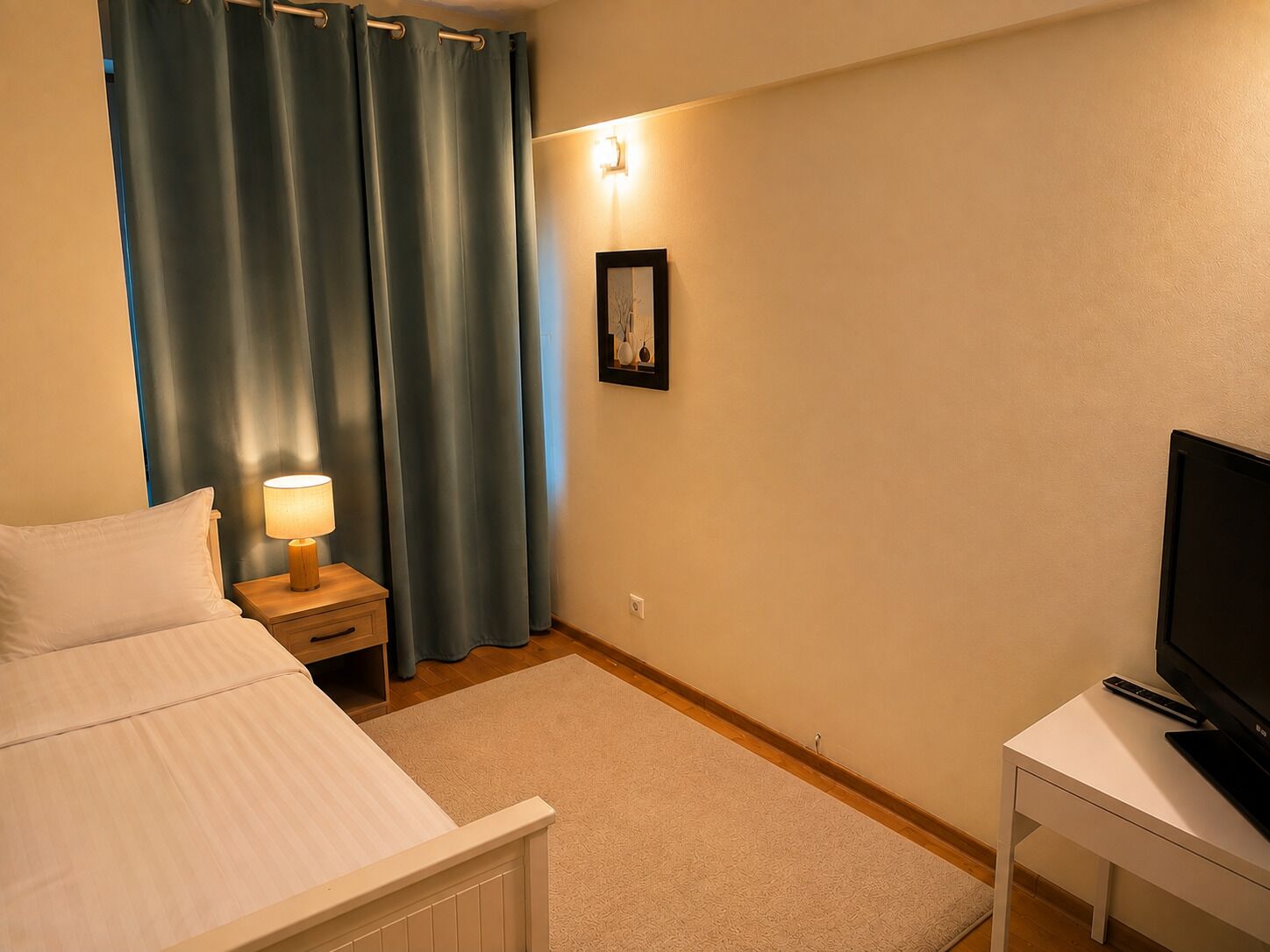 Una habitación de hotel con una cama, una mesita de noche con una lámpara, un televisor en un mueble bajo y un cuadro en la pared.