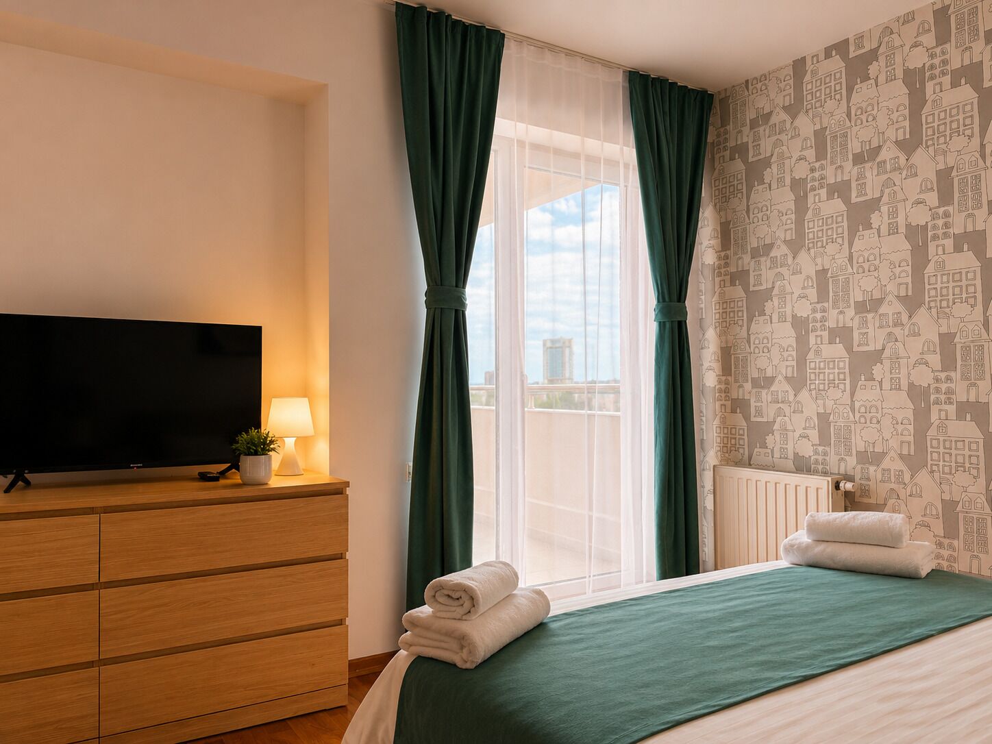 A hotel room with a bed, a TV on a wooden dresser, a window with green curtains, and a patterned wall.