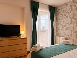 A hotel room with a bed, a TV on a wooden dresser, a window with green curtains, and a patterned wall. - Monte Carlo Palace Suites (Bucharest)