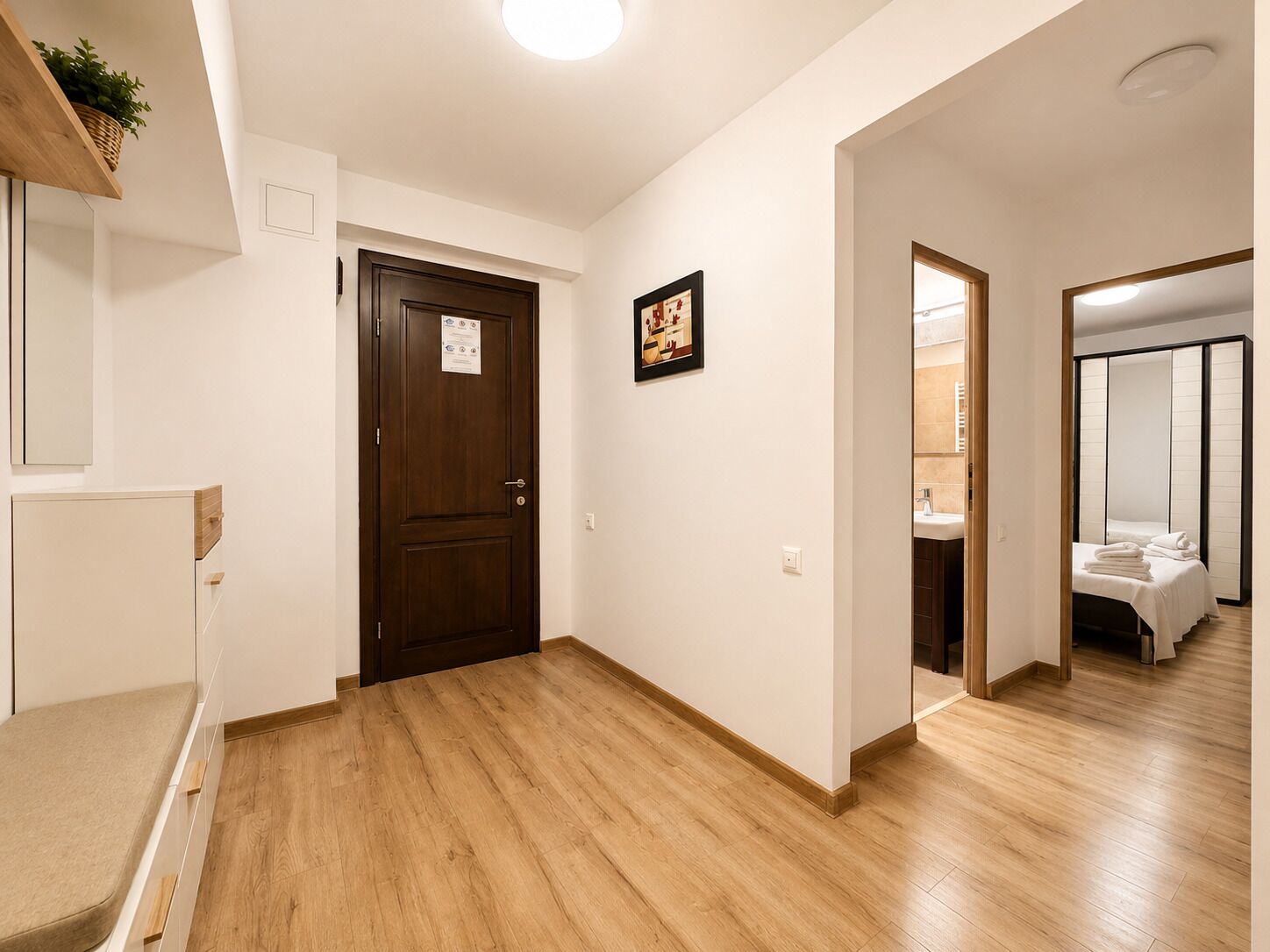 Quarto de hotel com piso de madeira, cama com roupa de cama branca, banheiro com pia e espelho, e uma porta com aviso.